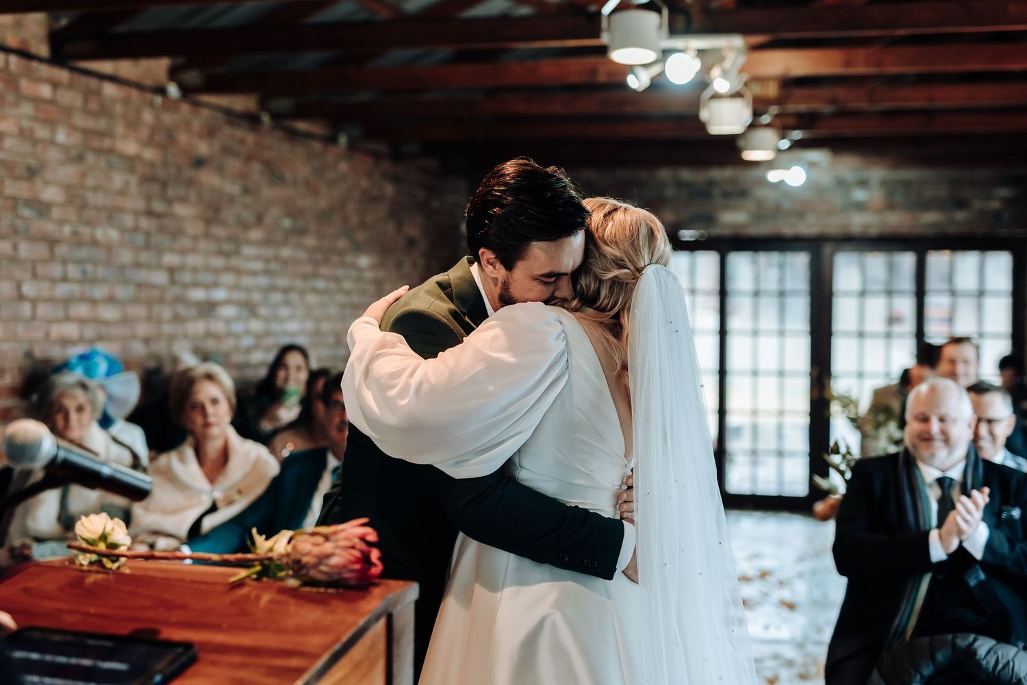 Couple shares their first kiss as newlyweds during reception with guests applauding in background.
