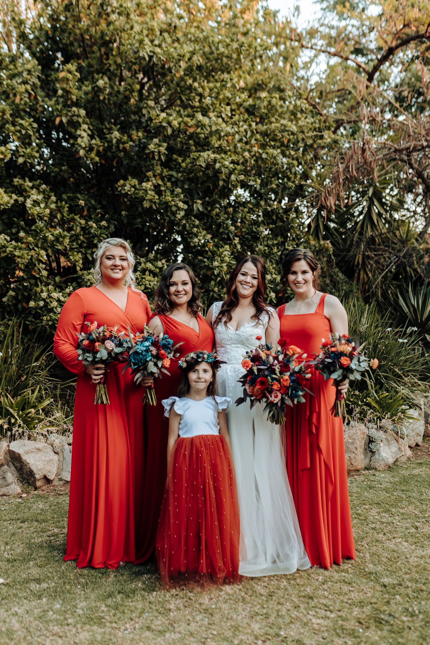 Wedding party with bridesmaids in coral red dresses and young flower girl in red tulle skirt holding floral bouquets.