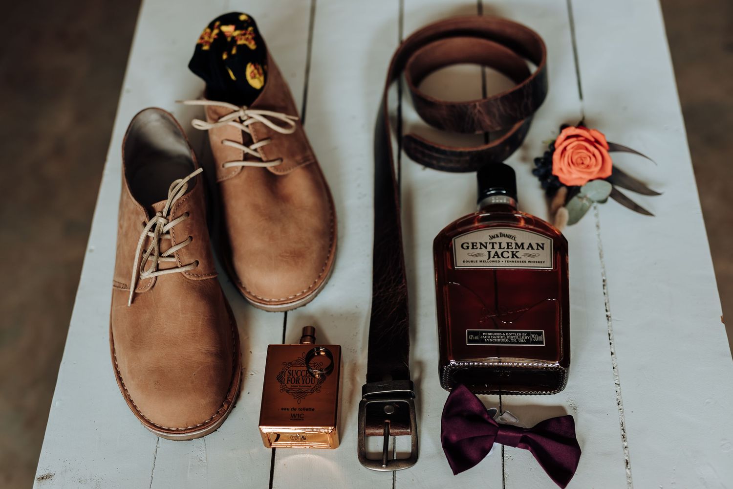 Wedding accessories including brown leather shoes, bow tie, belt, and bourbon bottle styled on white surface.