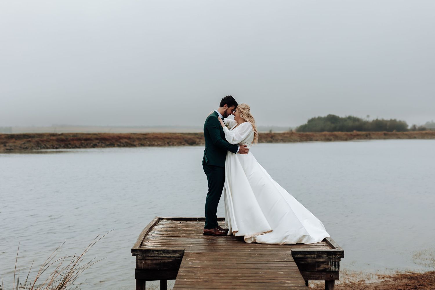 A couple shares a romantic moment in wedding attire while standing on a wooden dock over a serene lake.