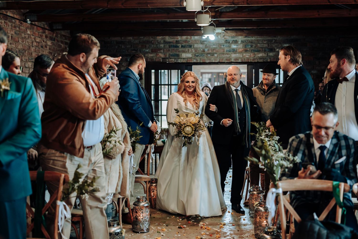 Wedding ceremony guests gathered in a rustic brick venue with exposed wooden beams.