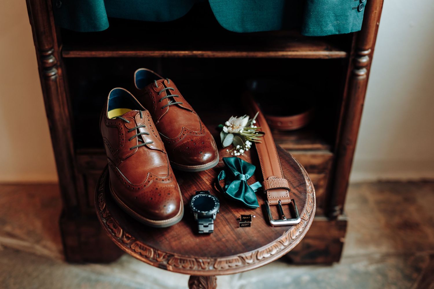 Close-up of brown leather dress shoes, belt, and accessories arranged on wooden table.