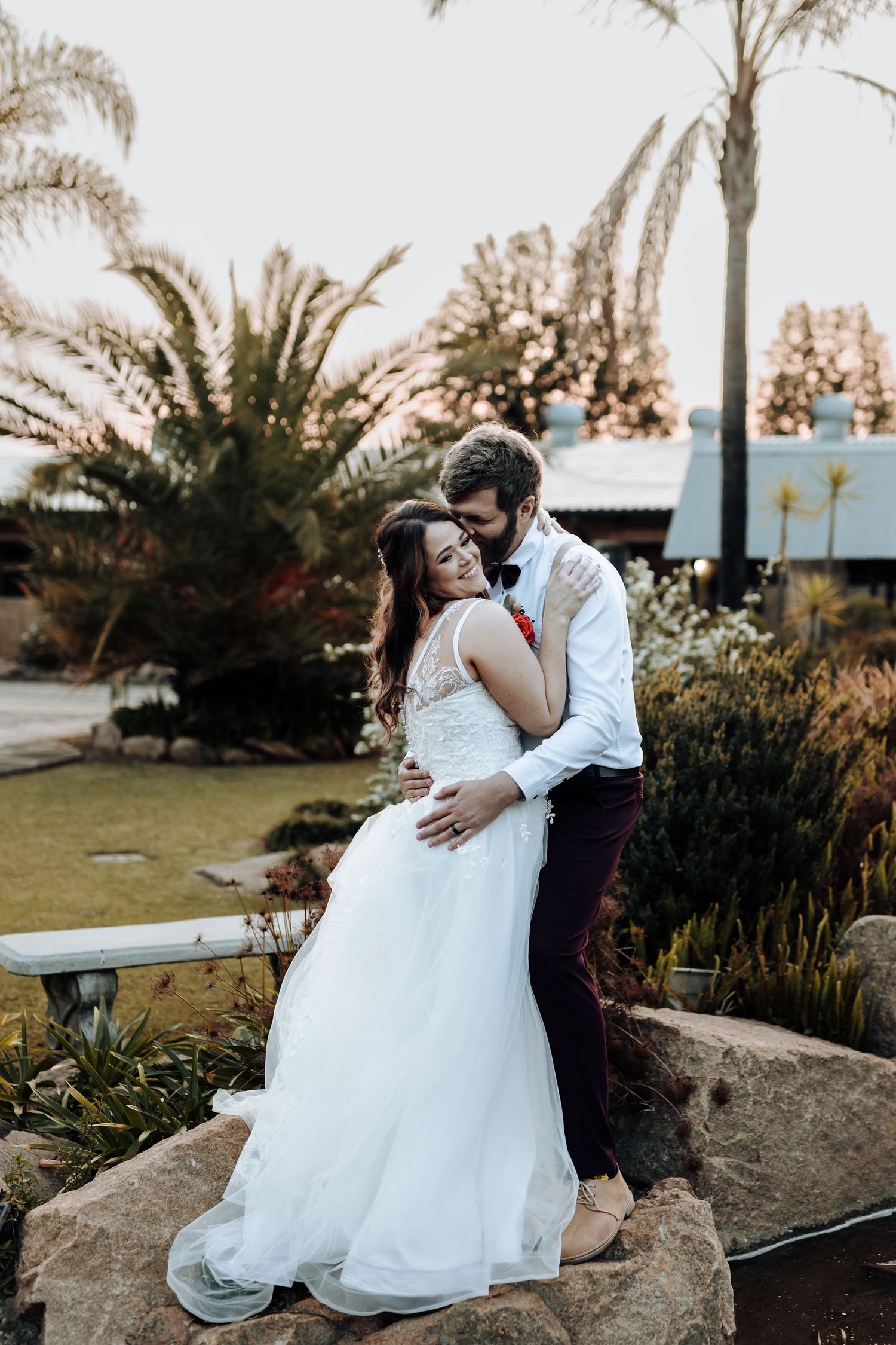 Elegant wedding portrait of couple standing on decorative rocks surrounded by lush garden.