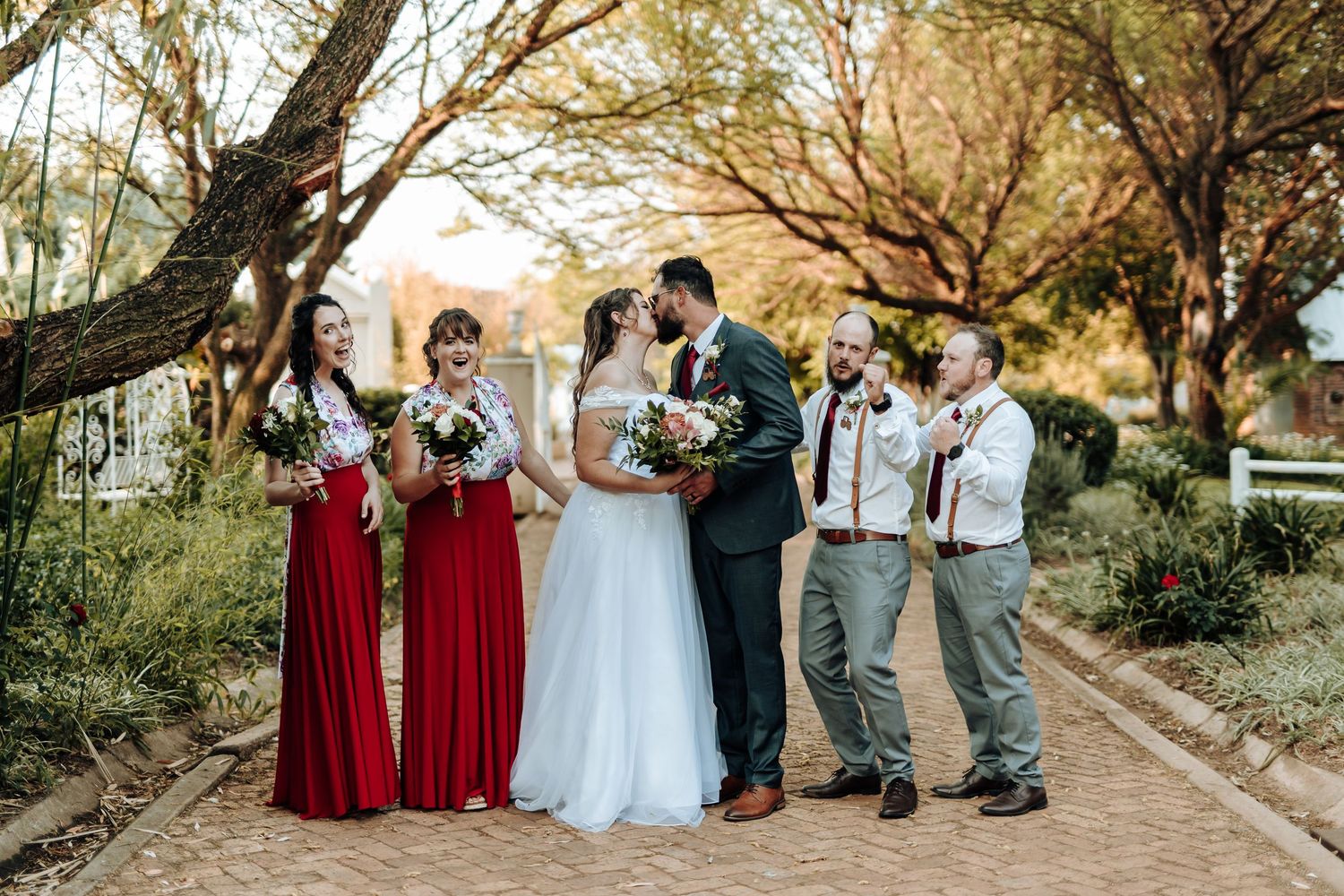 Wedding party members in red dresses and suspenders celebrate on an autumn path.