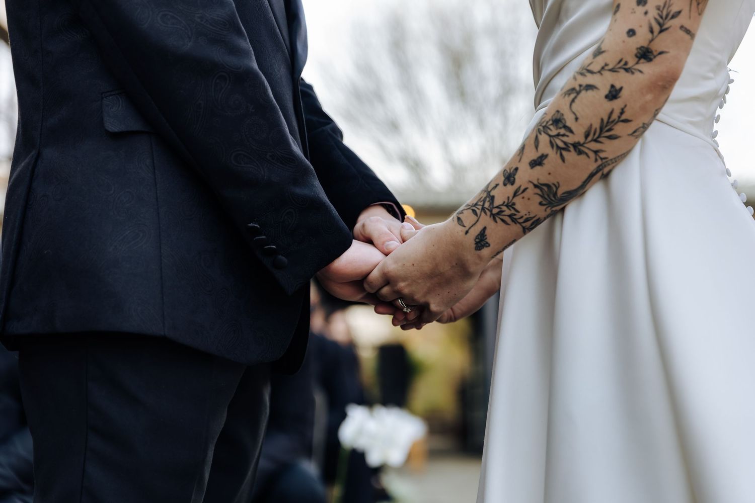 Close-up of couple holding hands, showcasing intricate floral tattoo sleeve against white wedding dress and dark suit.