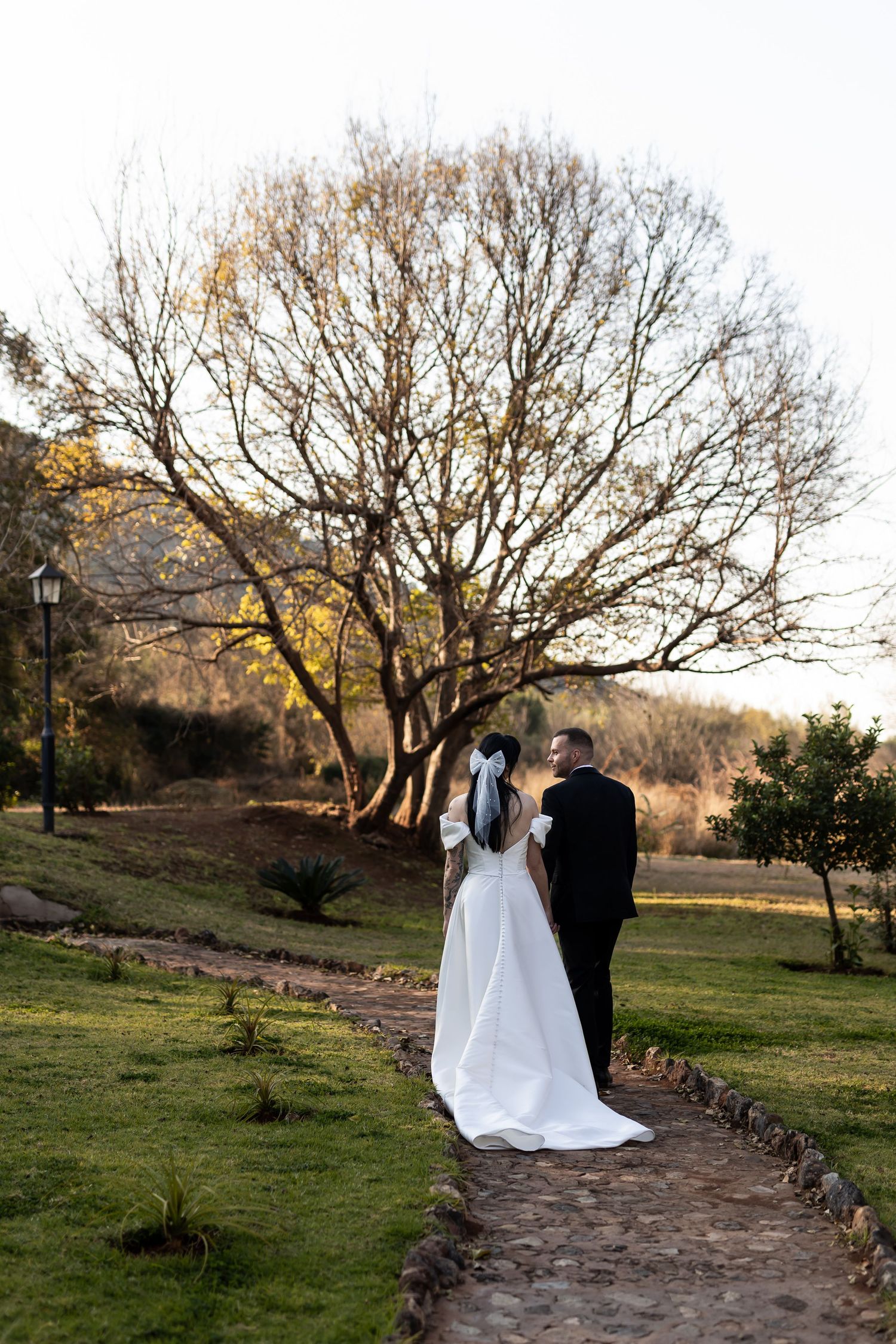 Couple in wedding attire stand on stone path under bare tree branches at sunset in countryside garden.