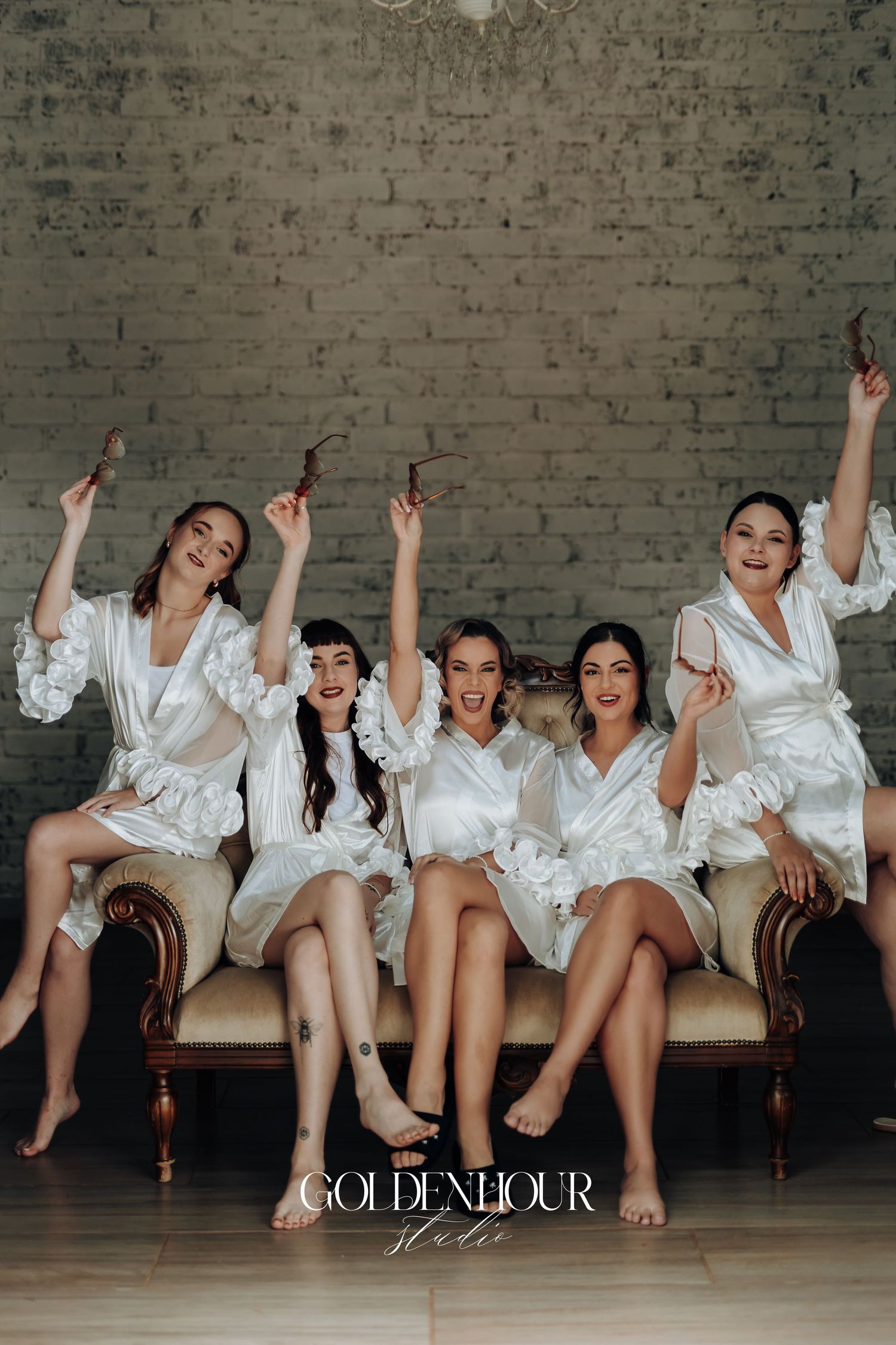 Wedding party wearing white robes poses playfully on vintage couch against brick wall.