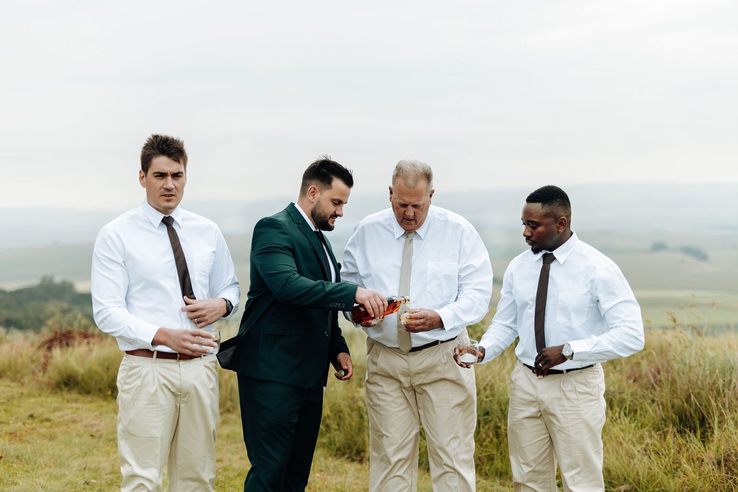 A group of well-dressed men in formal attire gather on a hilltop overlooking a scenic landscape while looking at a device.