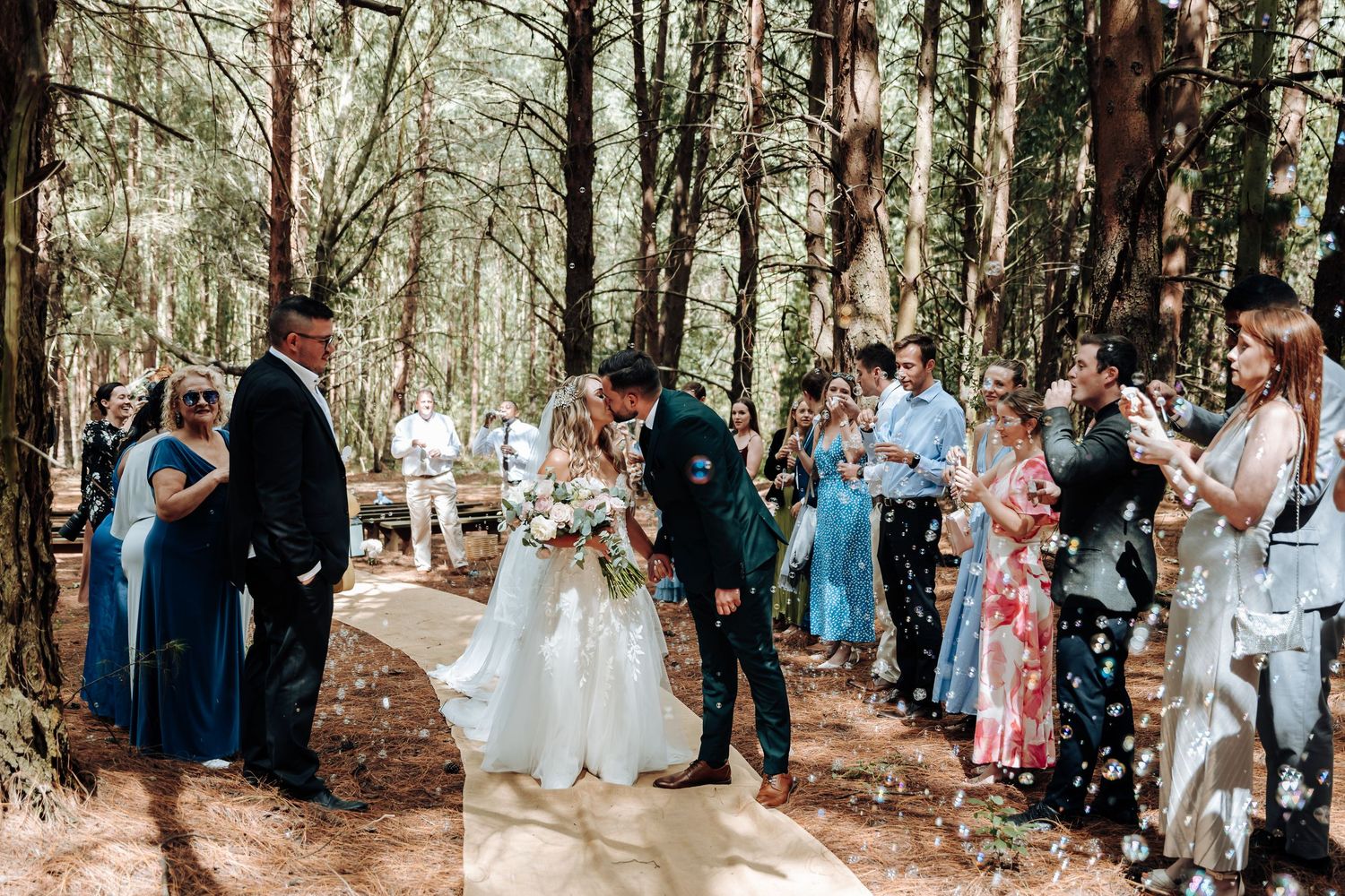 A wedding ceremony takes place in a serene forest setting with guests gathered along a wooded path.
