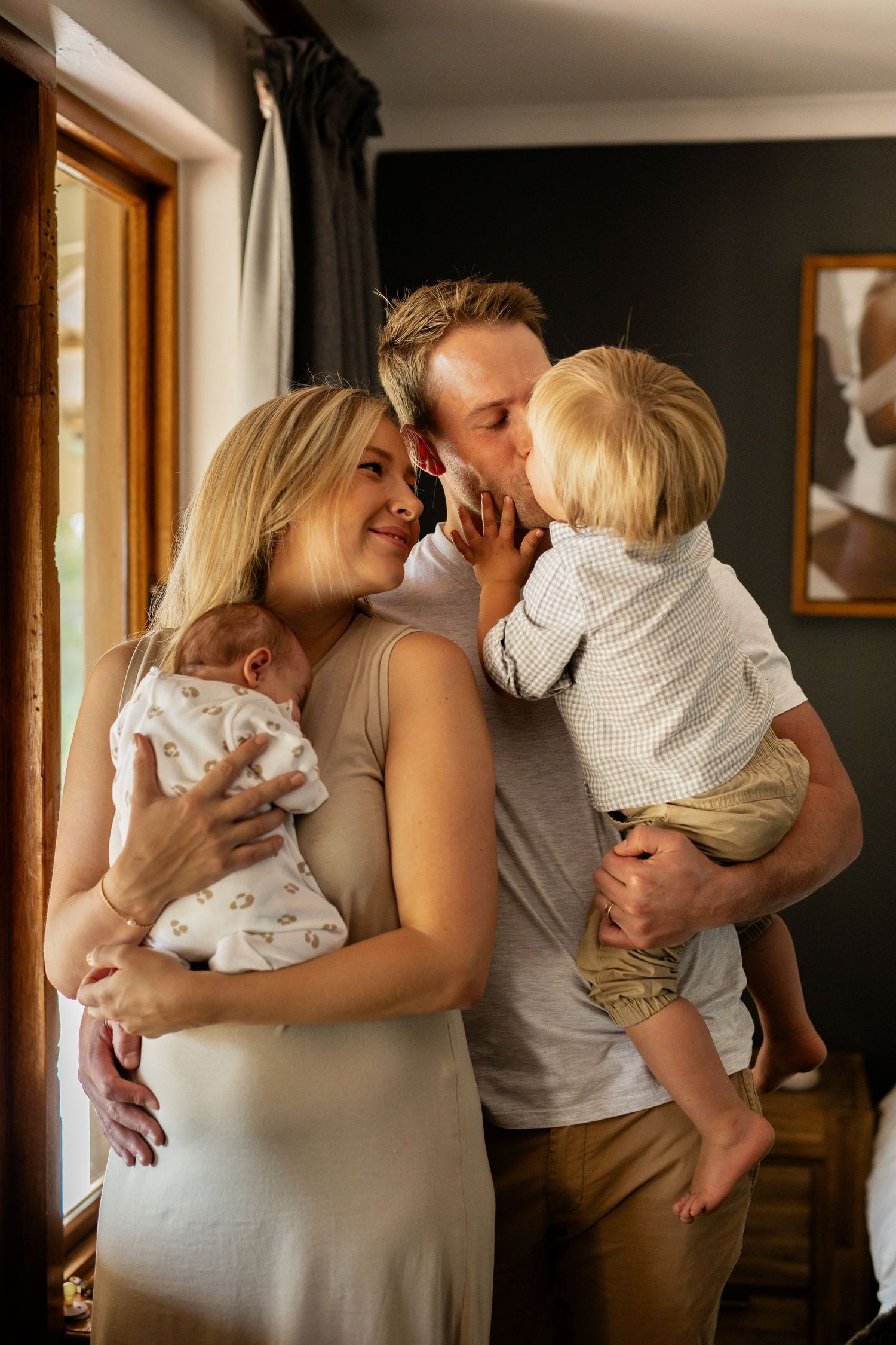 Parents share intimate moments with their newborn baby in a series of lifestyle photos by a sunlit window.