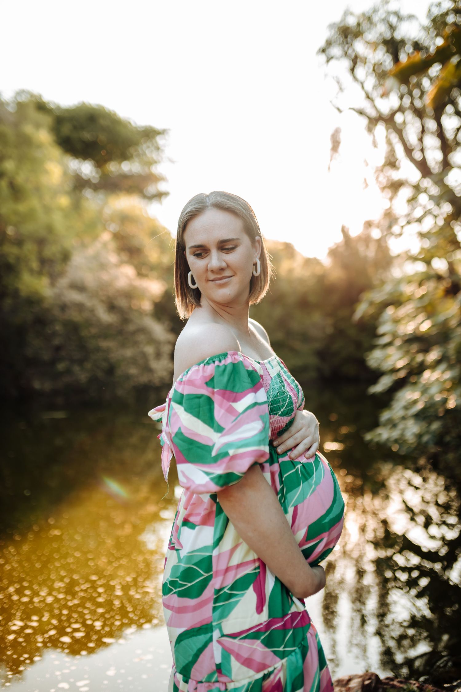 Expecting mother poses in floral pink and green dress by reflective pond during golden sunset.
