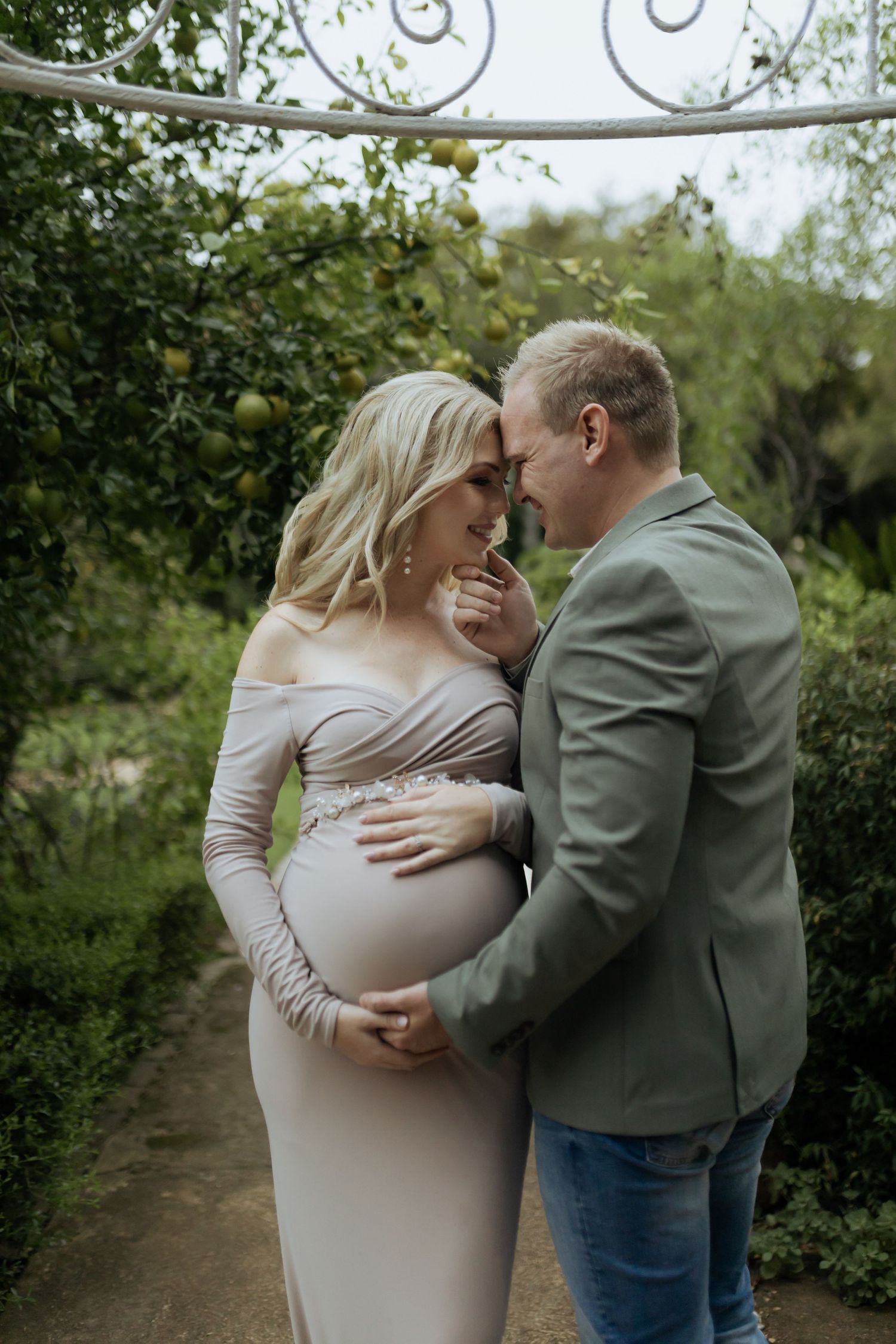 Expectant couple shares intimate moments in a garden setting wearing casual attire during maternity photoshoot.