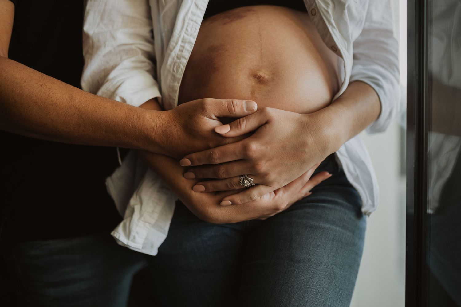 Close up of hands gently cradling a pregnant belly through an open white button-down shirt.
