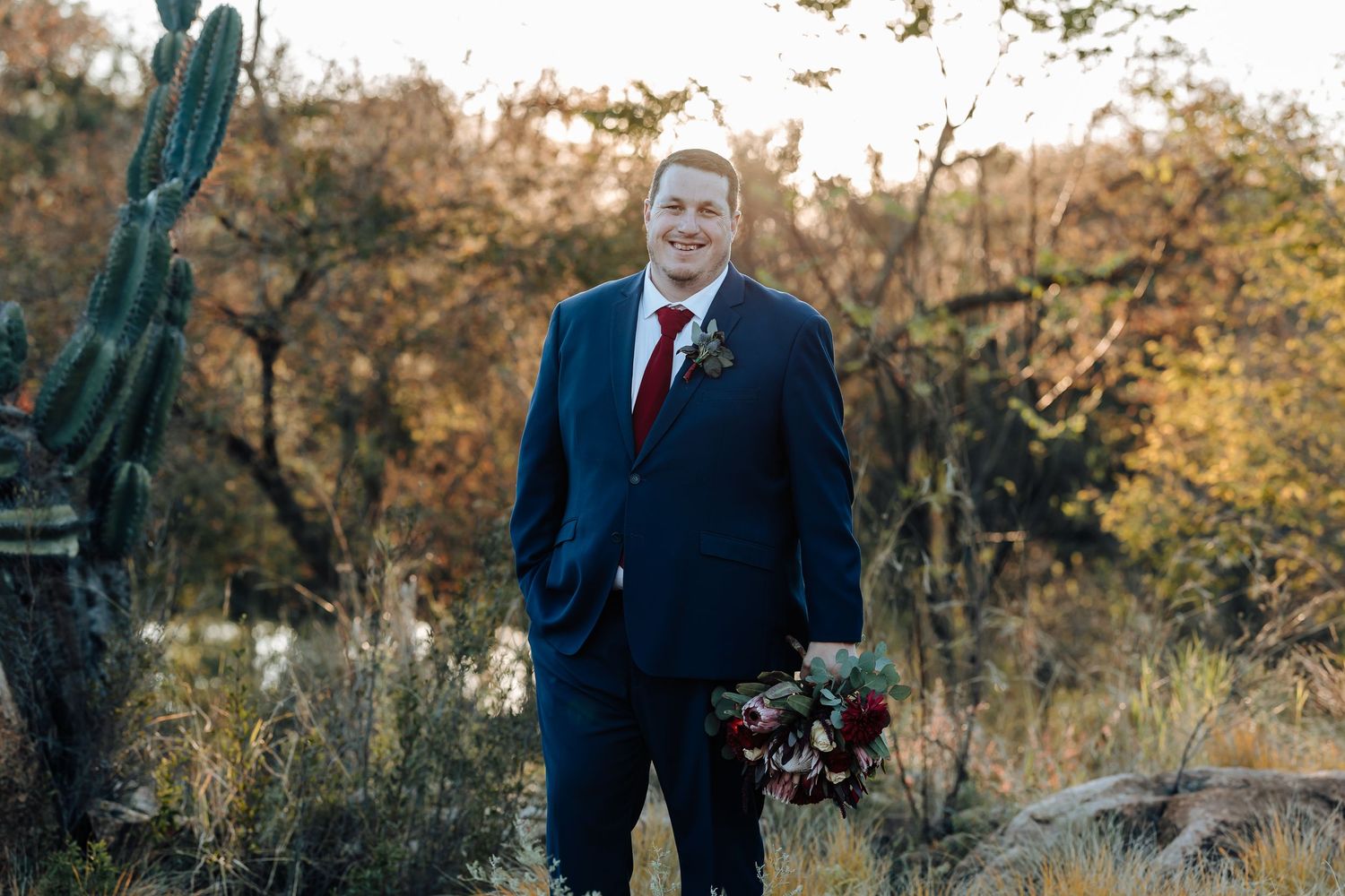 Groom in navy suit with red boutonniere holding bouquet in desert landscape with saguaro cactus.