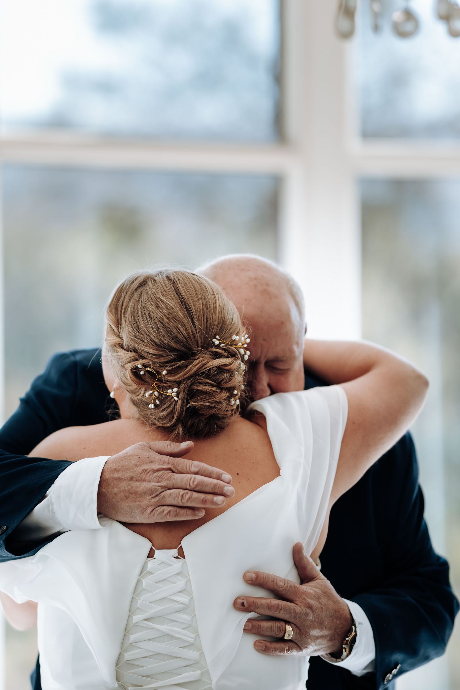 Bride in white dress with elegant updo hairstyle shares an emotional hug with older man in navy suit near bright window.