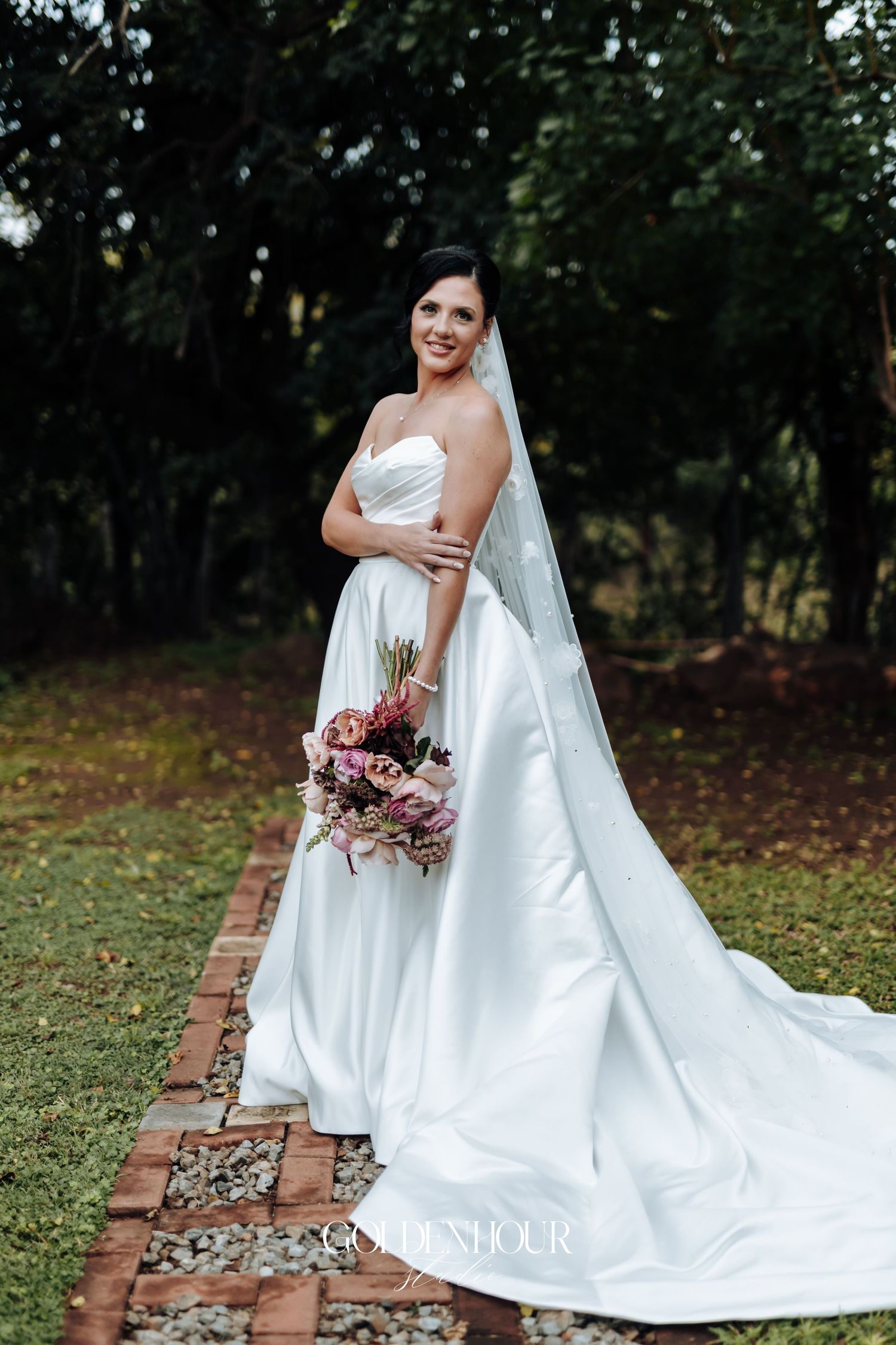 Beautiful bride in a strapless white satin gown with veil holding a pink bouquet outdoors.
