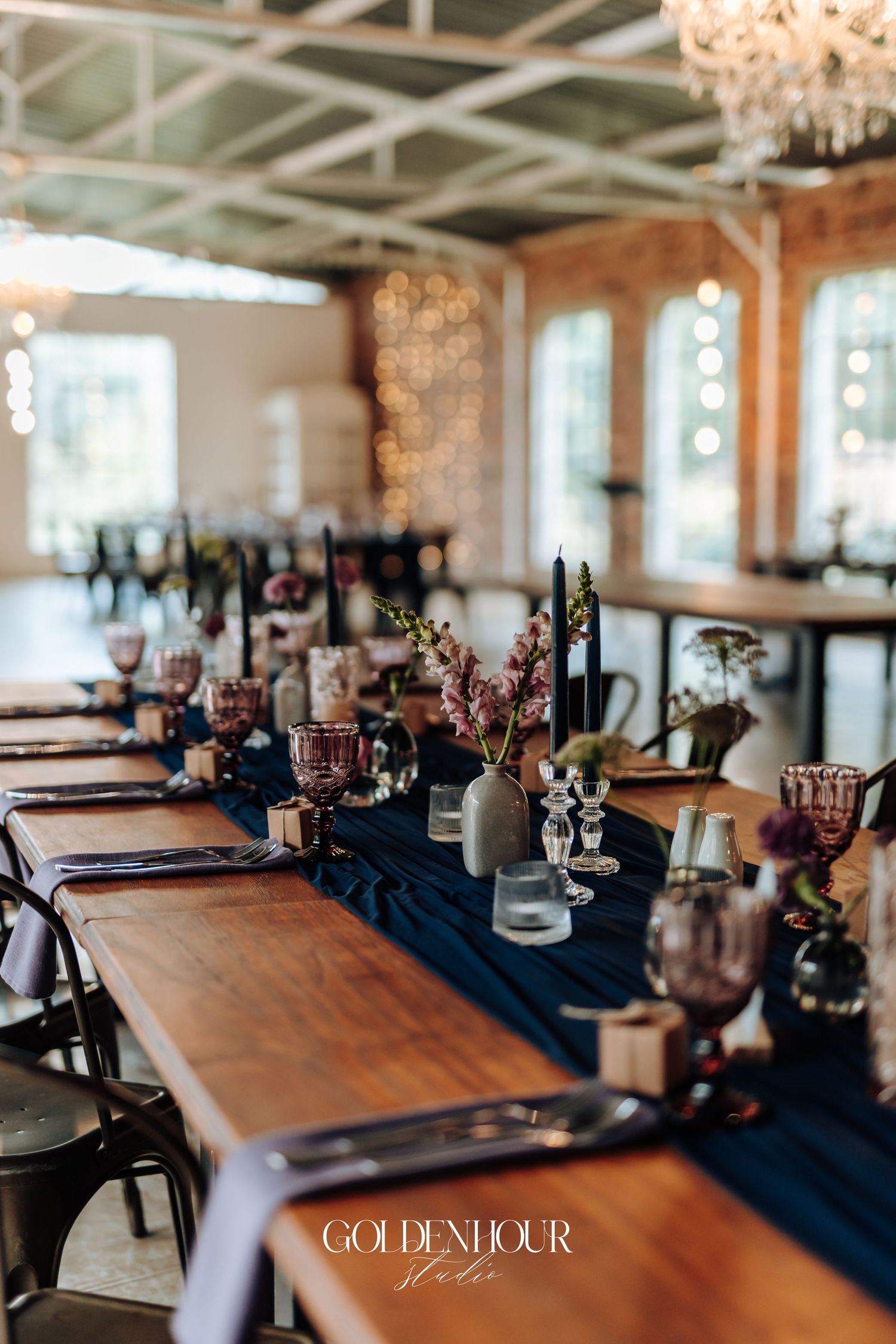 Elegant wedding reception table with navy blue runner, dark candles, pink florals, and vintage glassware in a rustic venue.