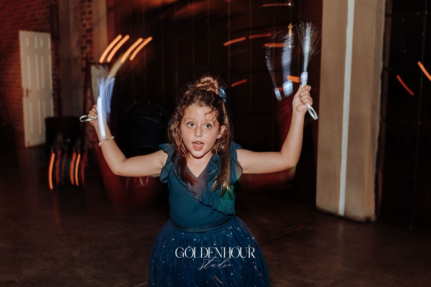 Excited young girl in teal dress holds champagne flutes above her head on a wedding dance floor.