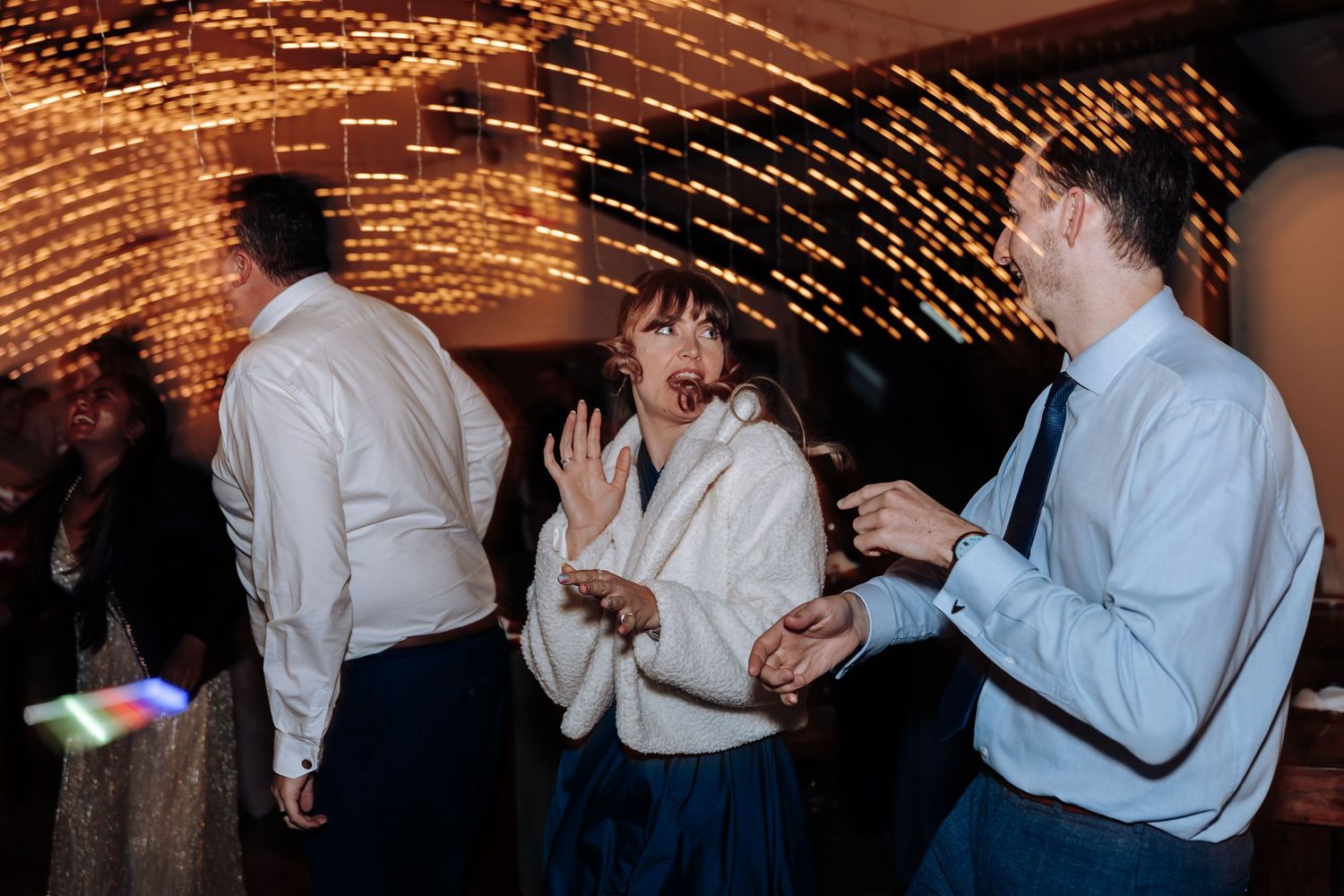 Guests dancing and laughing at a wedding reception with warm fairy lights glowing in the background.