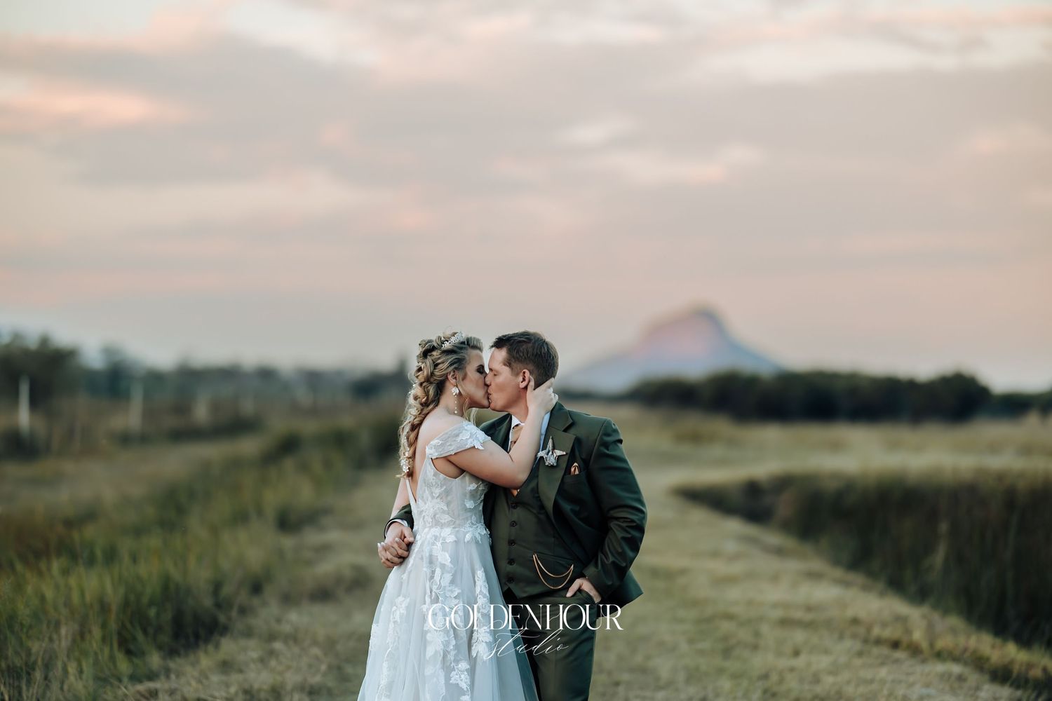 Romantic couple sharing a kiss in an open field at sunset with mountain backdrop.