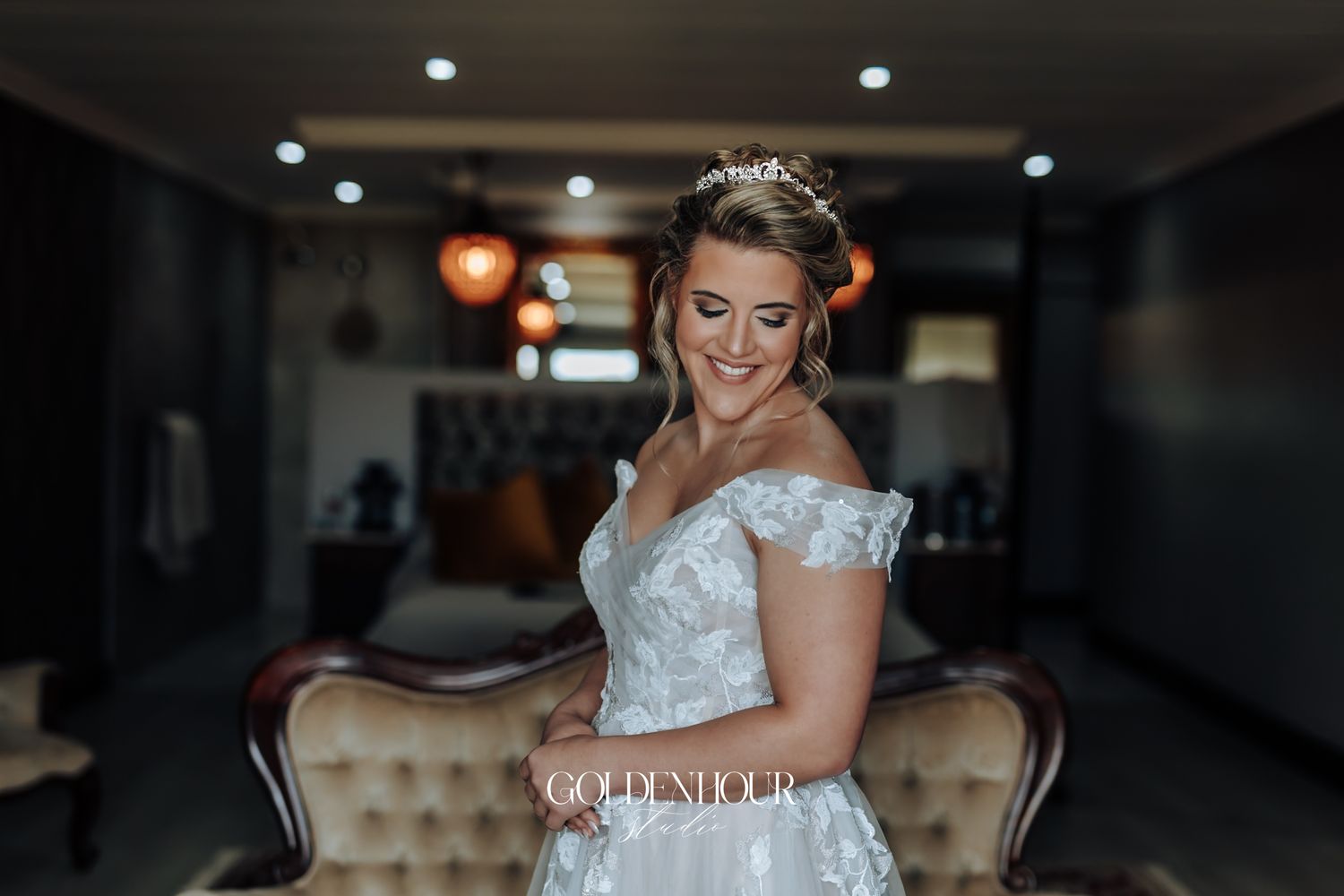 Bride in lace off-shoulder wedding dress wearing tiara smiling softly indoors.