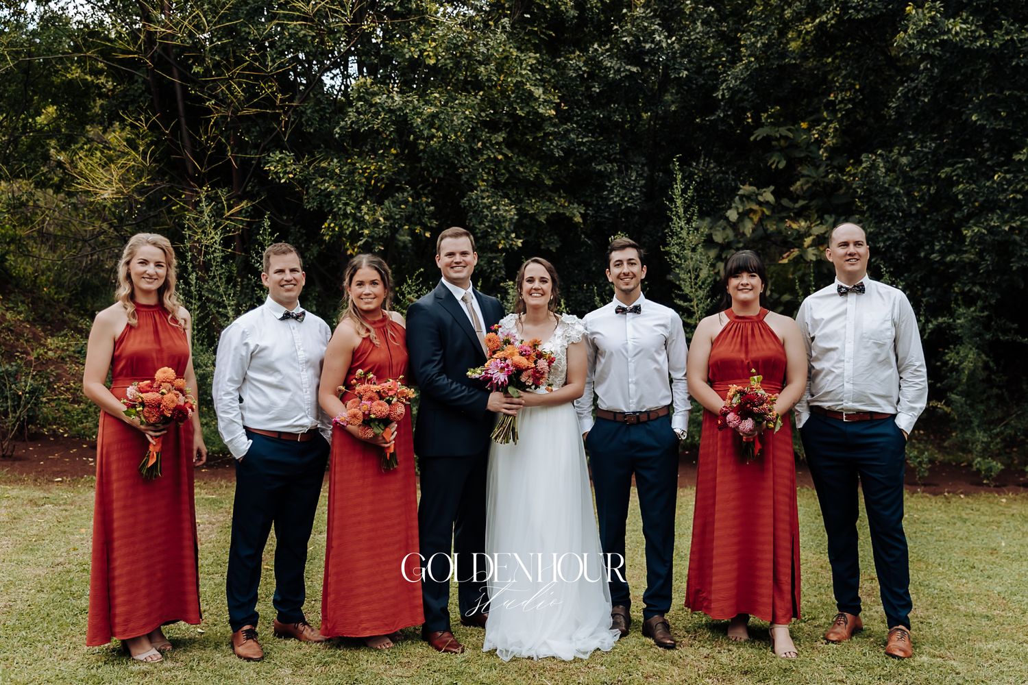 Bride and groom posing with full wedding party in red dresses and navy pants on green lawn.