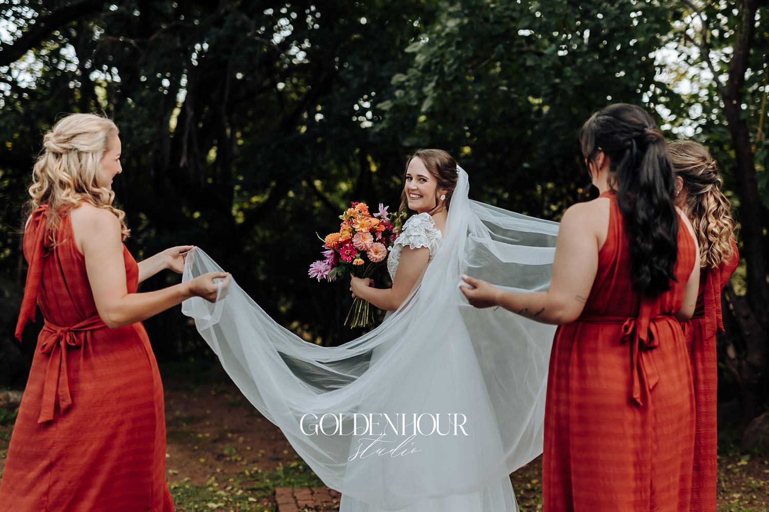 Bride in white gown and veil holding colorful bouquet assisted by bridesmaids in red dresses.