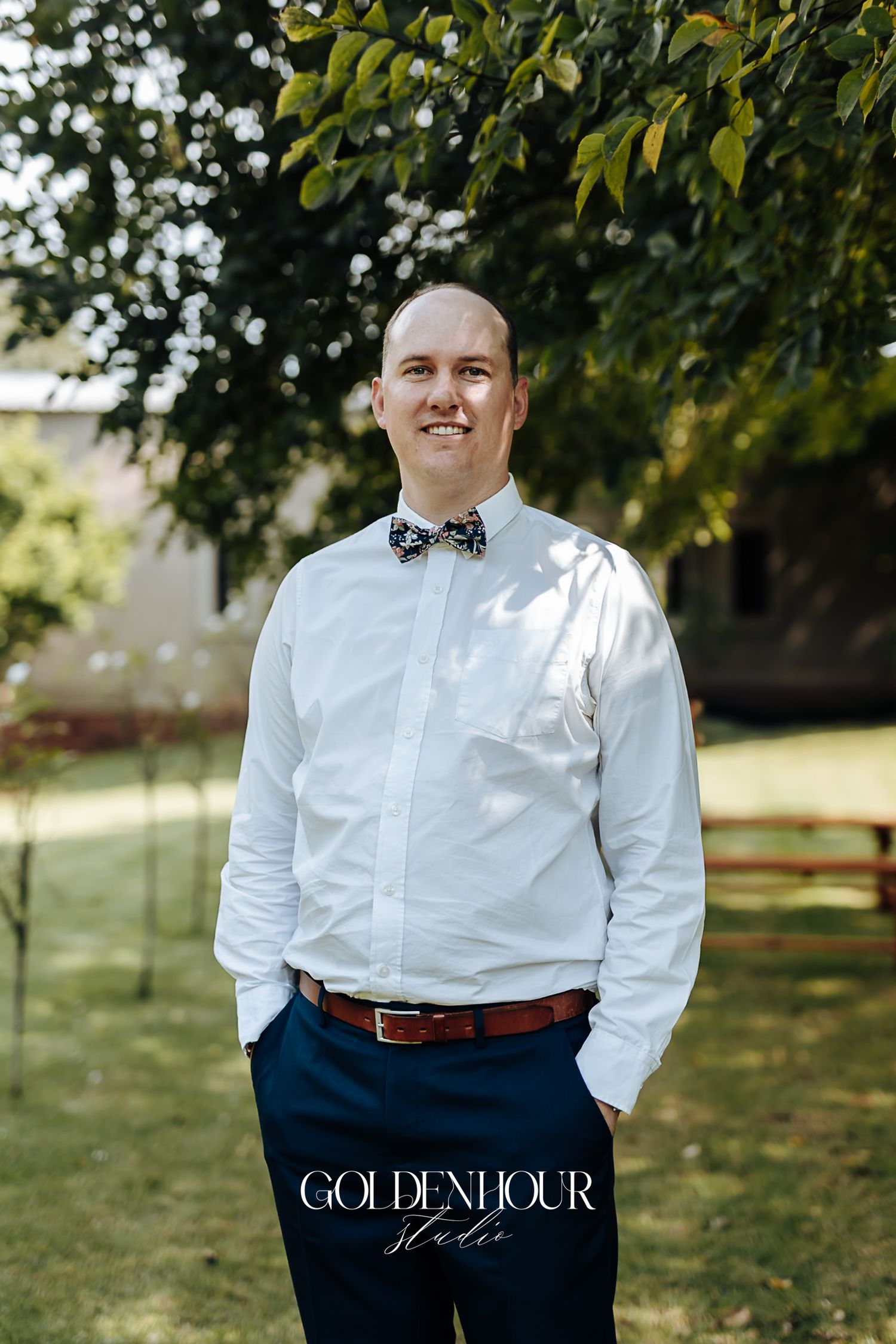 Groomsman smiling in a white shirt and floral bow tie in a garden wedding setting.