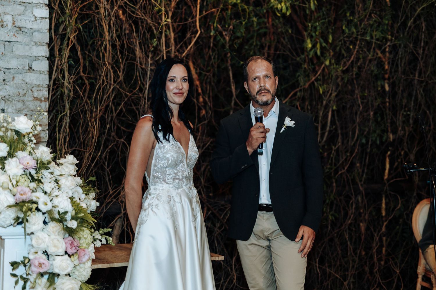 Bride in white gown and groom in navy blazer smiling during wedding reception speech near floral arrangements.
