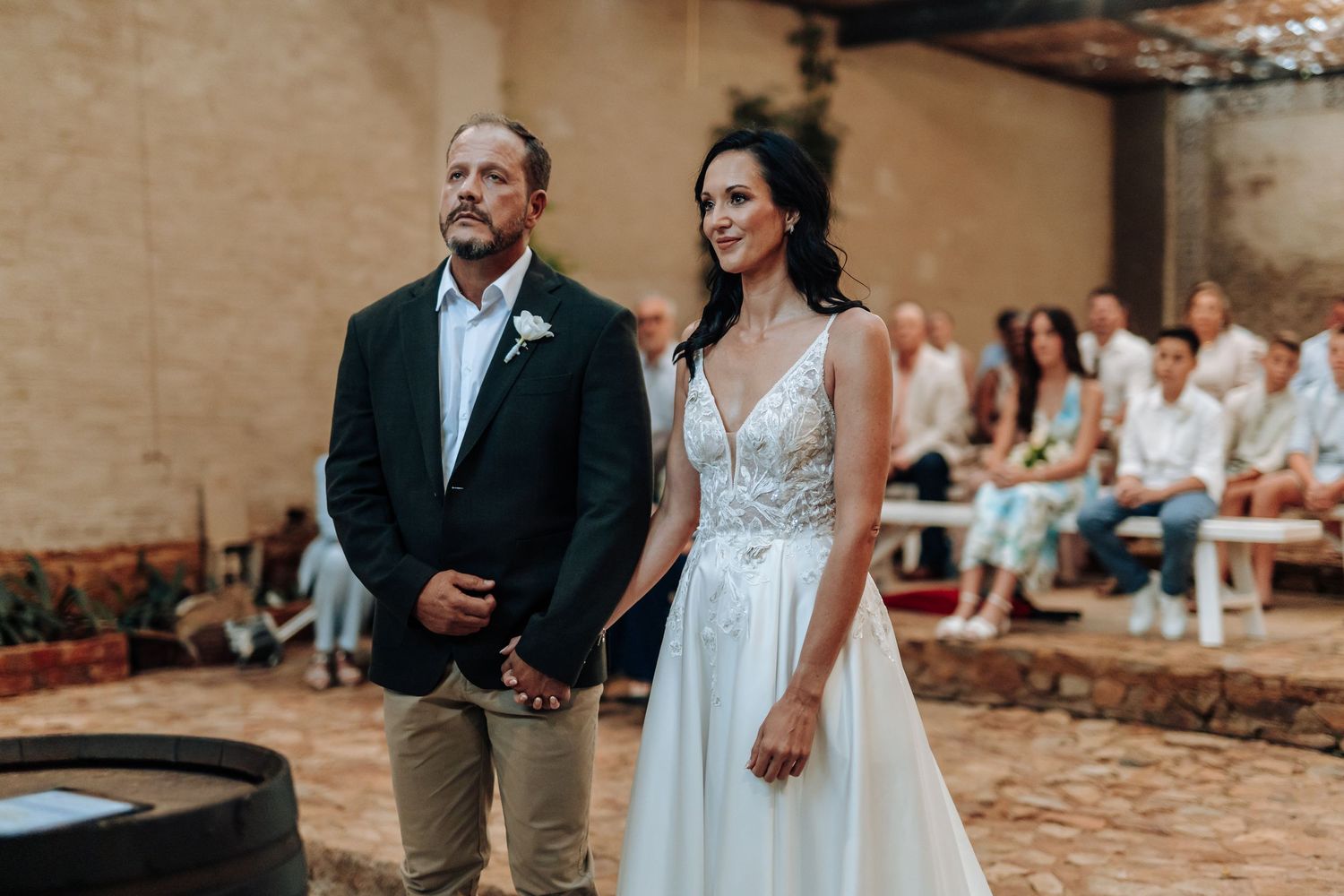 Bride in white gown walks down aisle with escort at rustic outdoor wedding ceremony with seated guests.