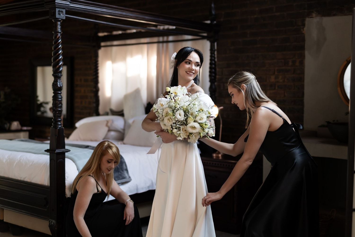 Bridesmaids in black dresses help adjust a bride's elegant white gown while holding a stunning white floral bouquet.
