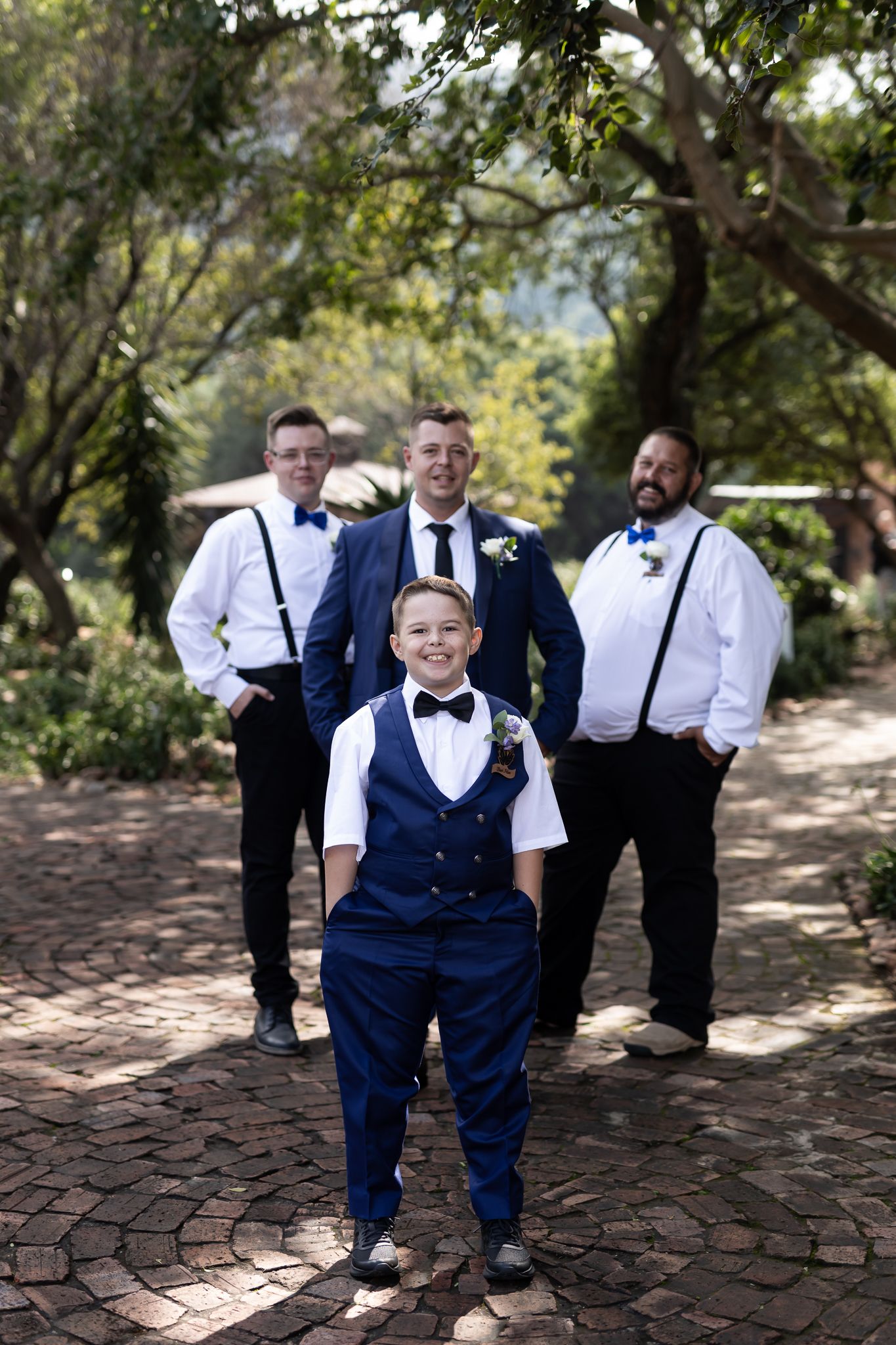 Wedding party members in coordinated navy and white attire pose on a brick pathway surrounded by trees.