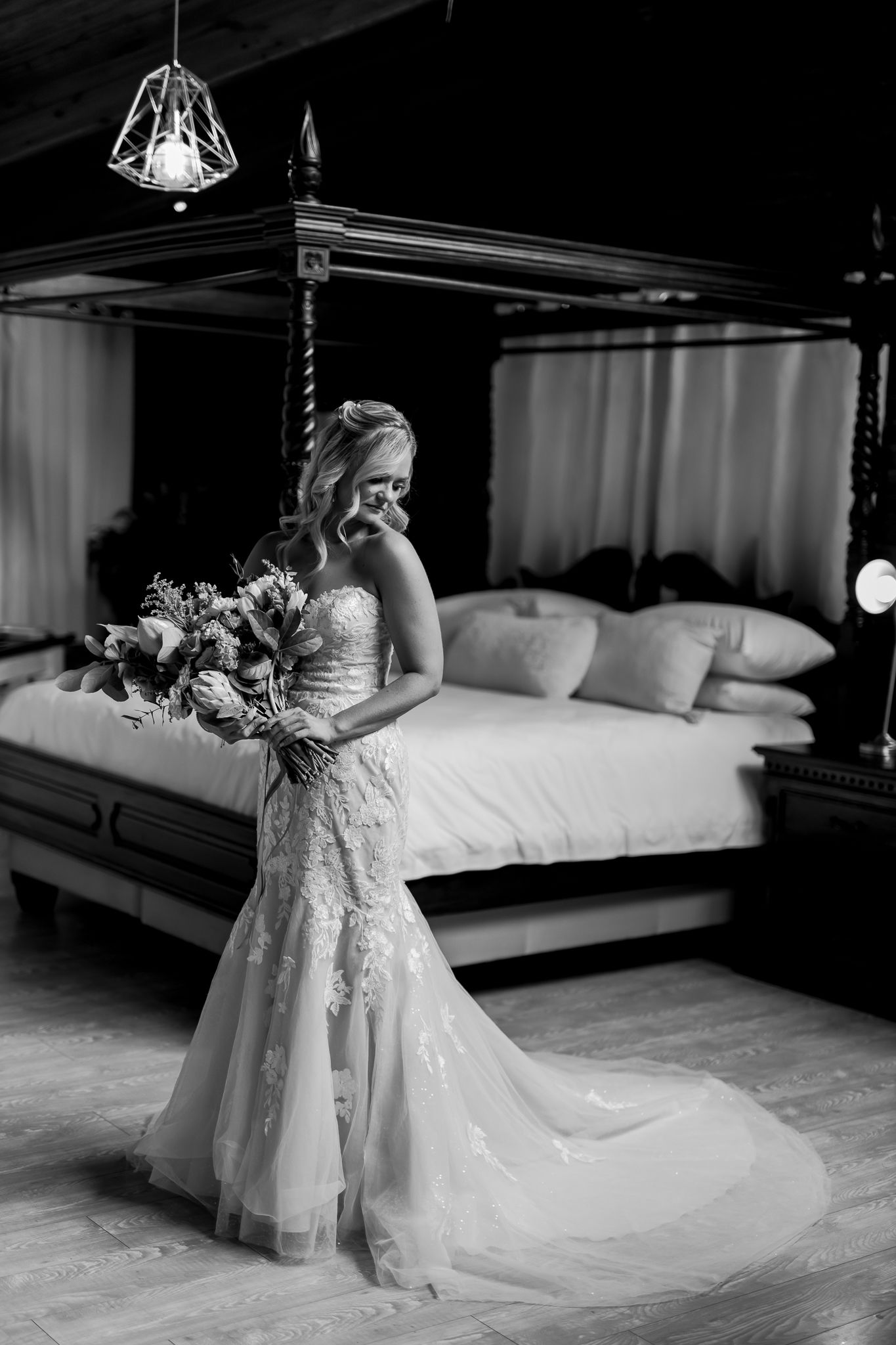 Black and white photo of a bride in a flowing lace gown posing in front of a canopy bed.