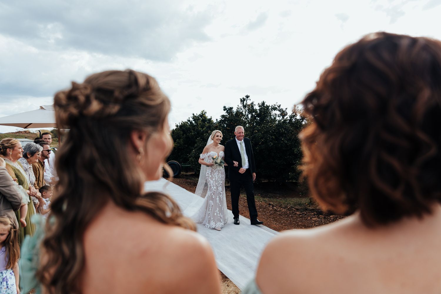Wedding guests watching the ceremony at an outdoor rustic venue with white fabric decor under cloudy skies.