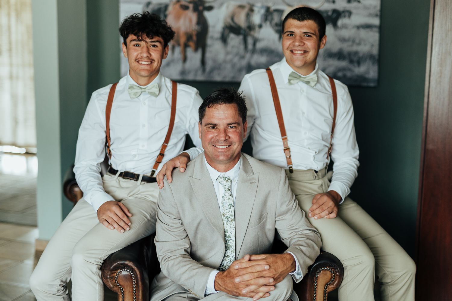 Groomsmen in light colored suits with suspenders and bowties pose together on a leather chair.