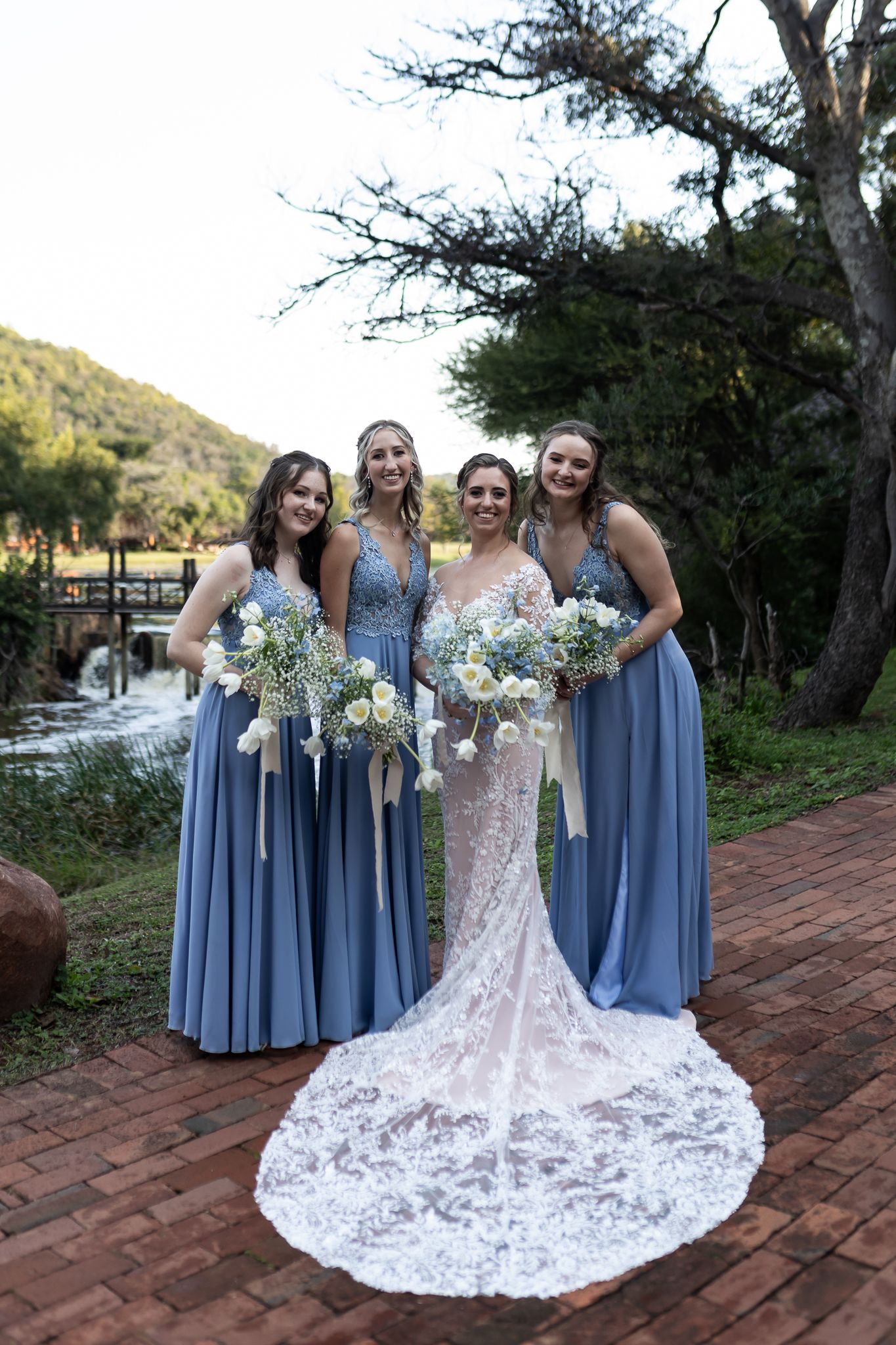 Bridal party poses on brick pathway showcasing elegant lace wedding gown with long train.
