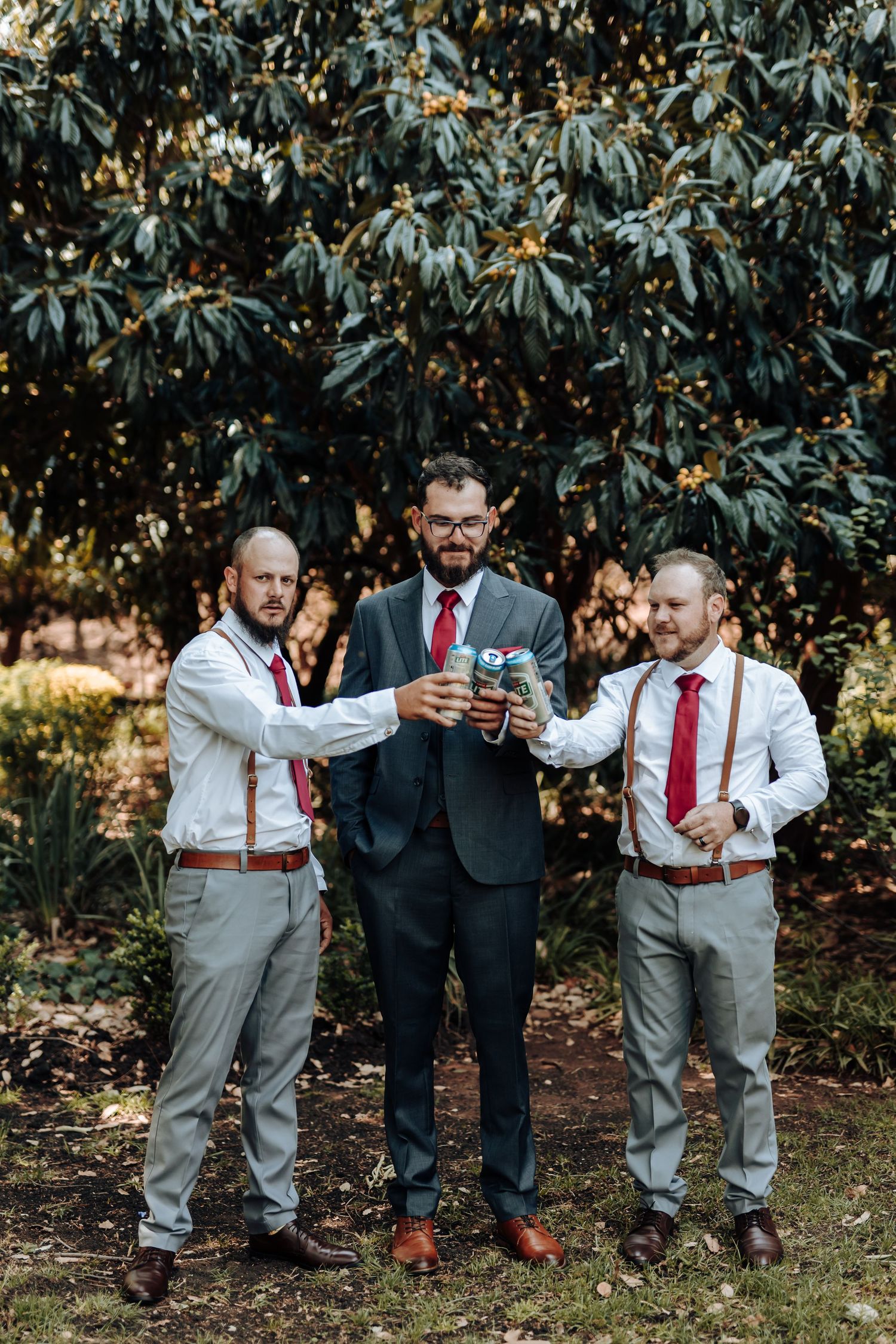 Groomsmen toast with drinks under a leafy tree during wedding celebrations.