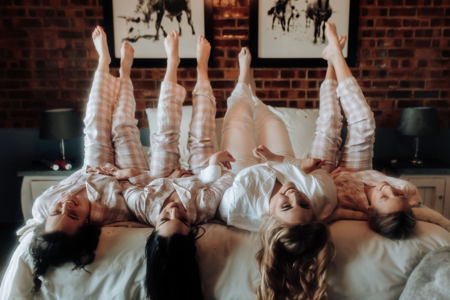 Friends in white outfits lay head-to-head in a circle on a bed, raising their legs in a playful pose.