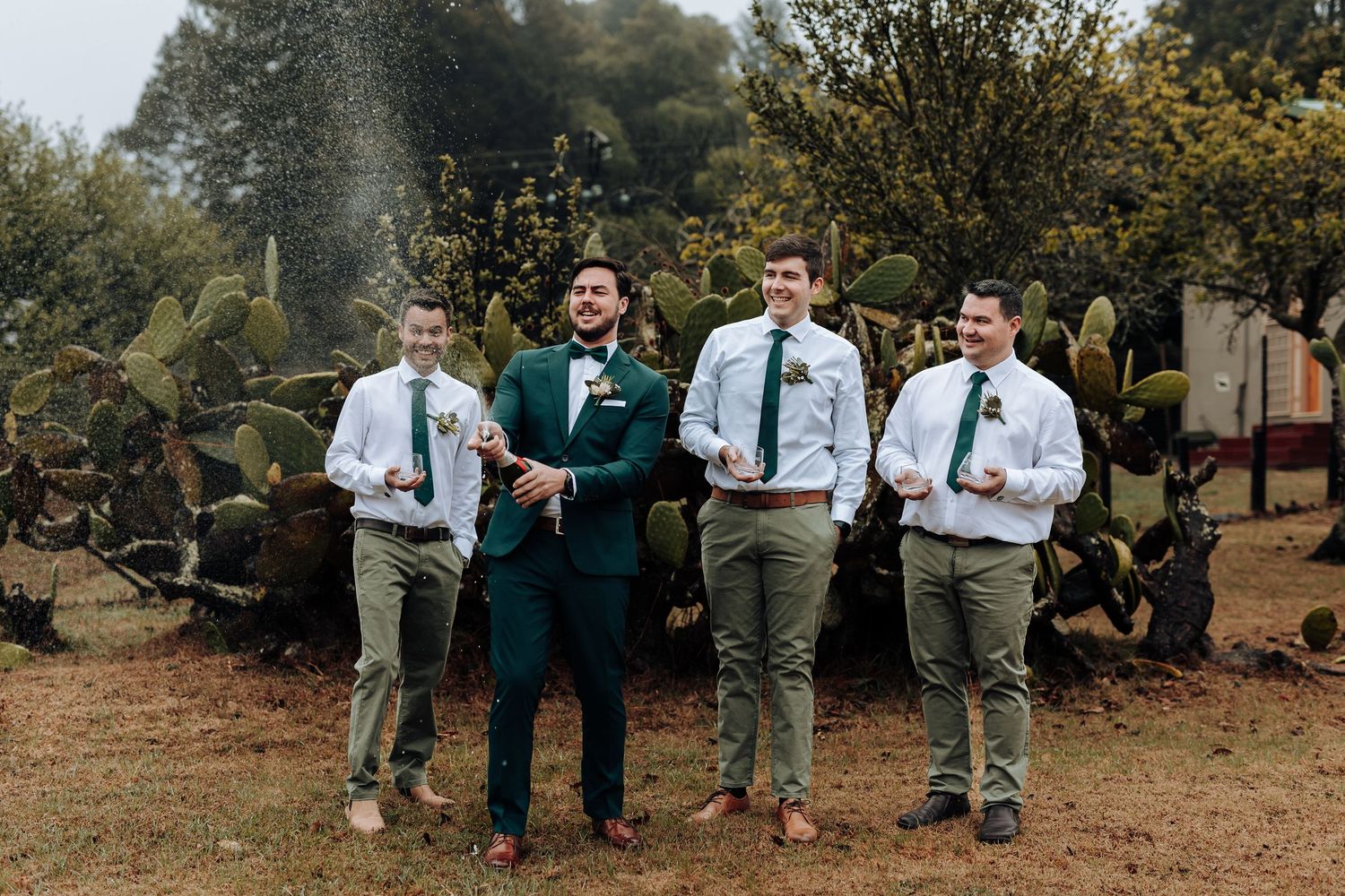 A groom in emerald suit stands with groomsmen in white shirts and khaki pants against a rustic outdoor backdrop.