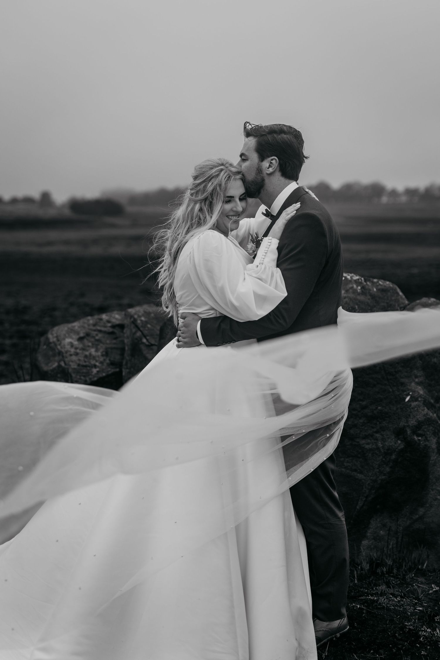 Black and white wedding photo captures couple's romantic embrace on rocky terrain with billowing veil.
