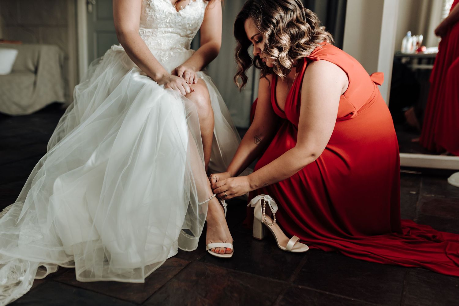 Bridesmaid assists with the bride's shoes during wedding preparations.