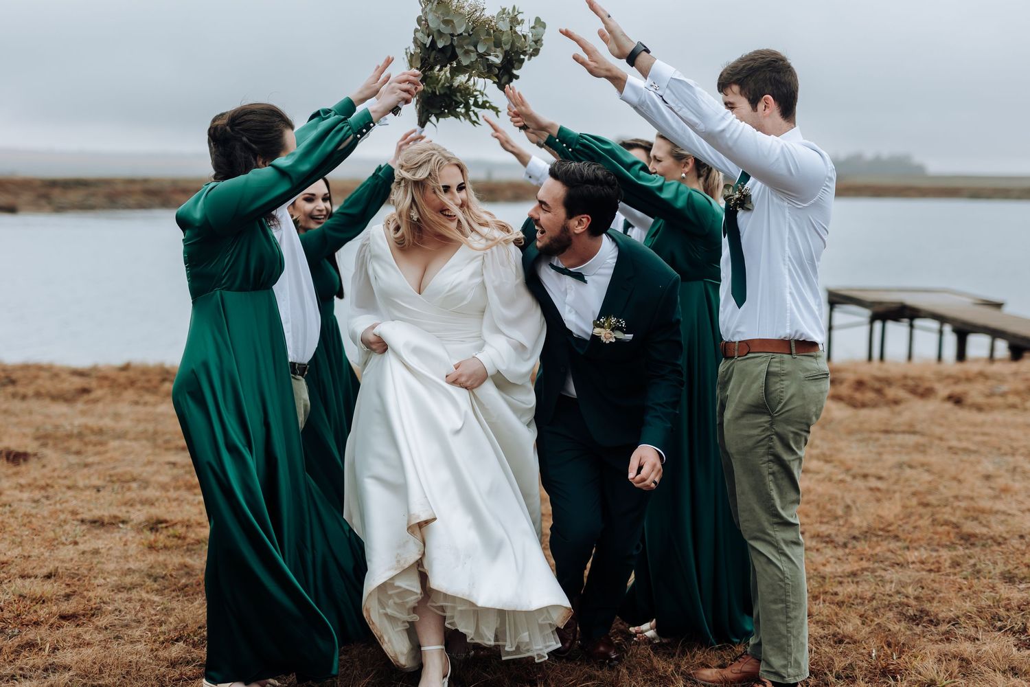 Bridal party creates playful arch for couple to walk under near water.