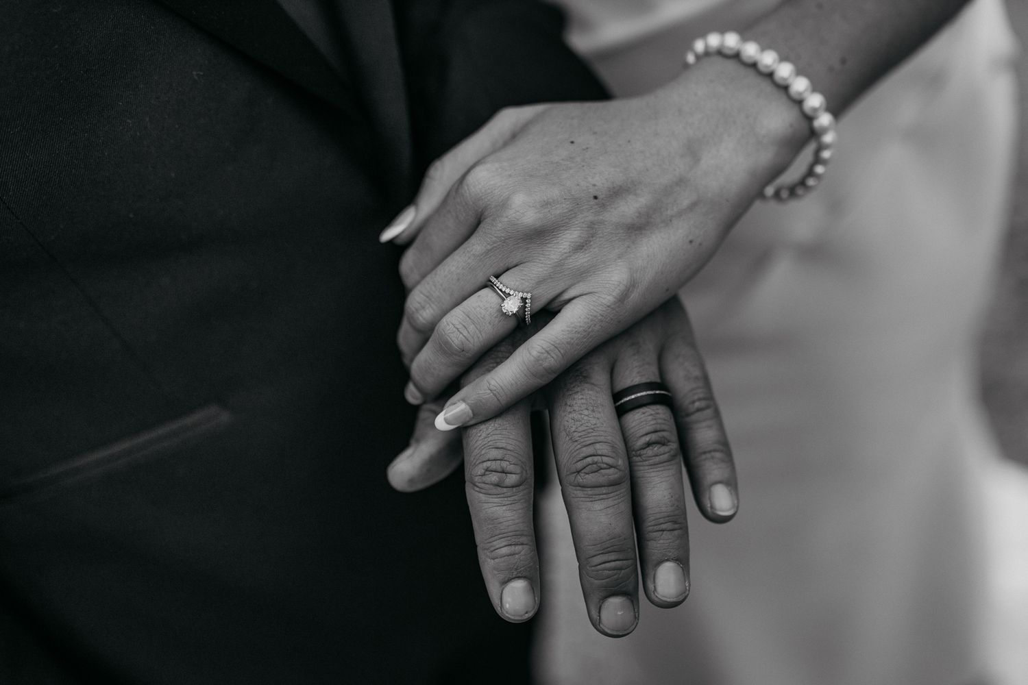 Close-up black and white photo showcasing wedding rings and clasped hands with delicate jewelry.
