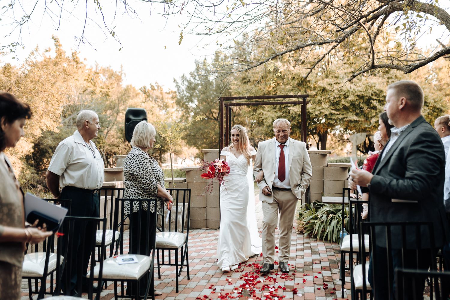 Outdoor wedding ceremony with red petals scattered along the aisle under an autumn arbor.