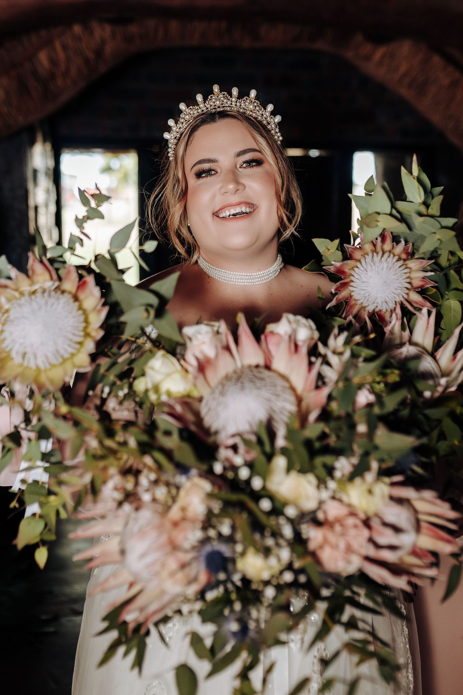 Bride in crystal crown poses with elegant bouquet featuring protea flowers and greenery in romantic wedding portrait.