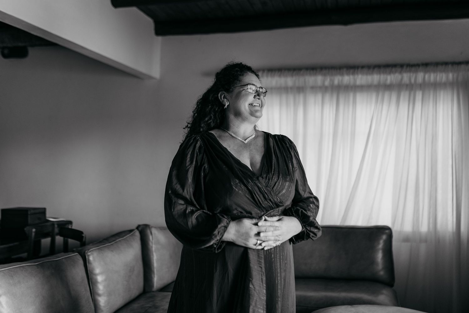 Black and white photograph of a person in a dark dress standing thoughtfully by a couch near curtained windows.