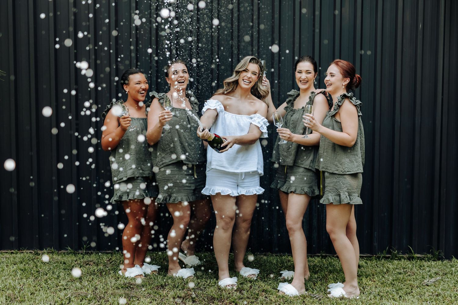 A group celebrates outdoors with bubbles while wearing coordinated olive green and white outfits.