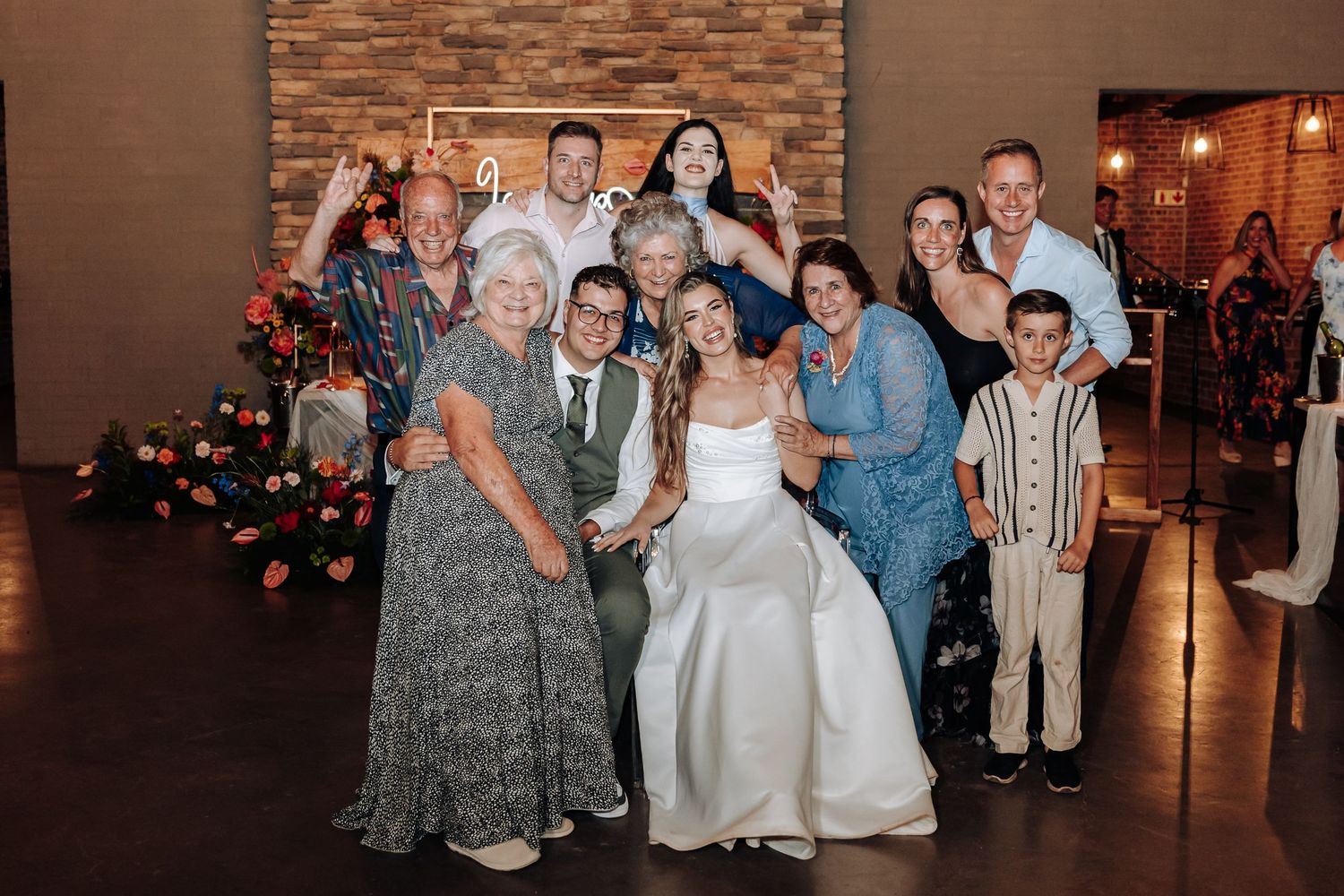Large group of wedding guests gathered for photos in front of a rustic brick wall.