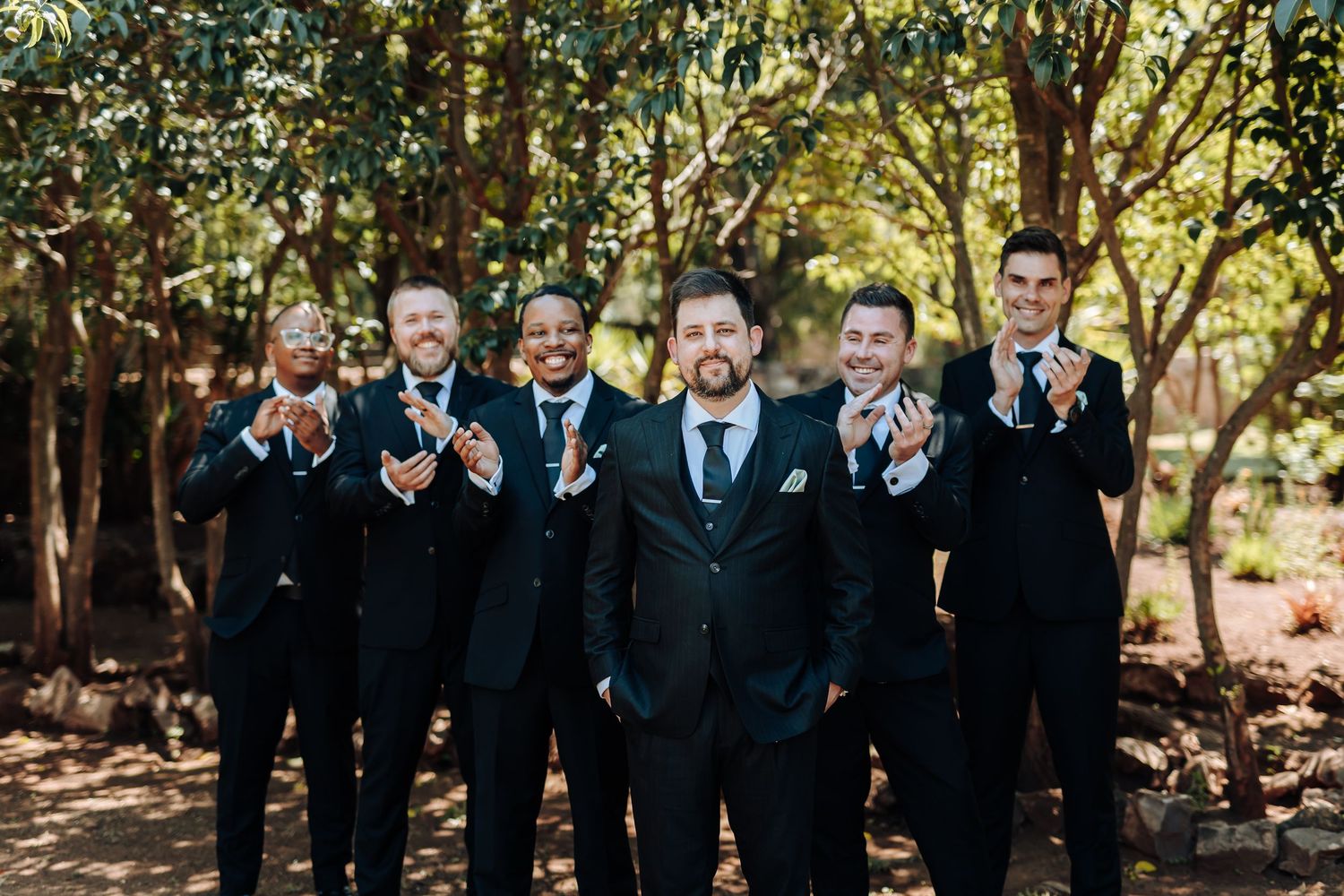 Wedding party members in formal black attire gather among trees in natural outdoor lighting.