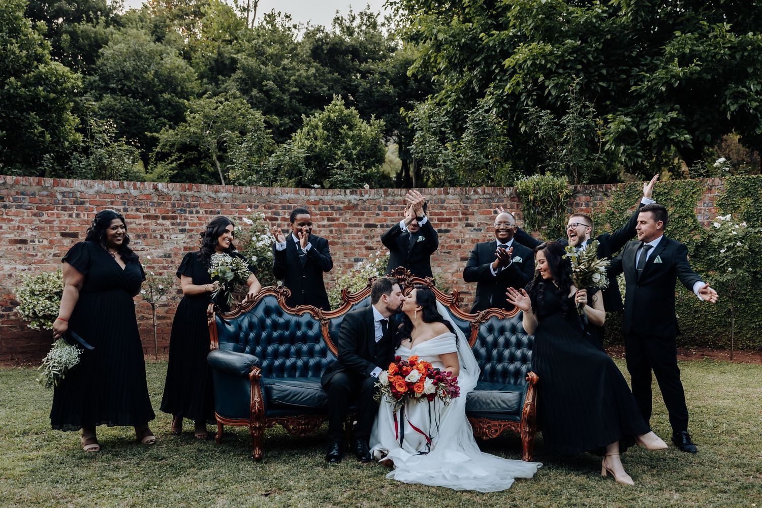 Wedding party posing against brick wall in formal attire with bridesmaids in black dresses.